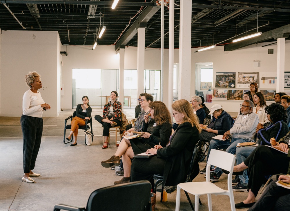 A woman stands speaking to a large seated crowd in a sparsely decorated room with white wall