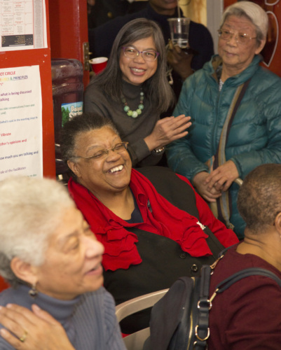 A group of smiling older adults of mixed races gather in a room, some are sitting some are standing
