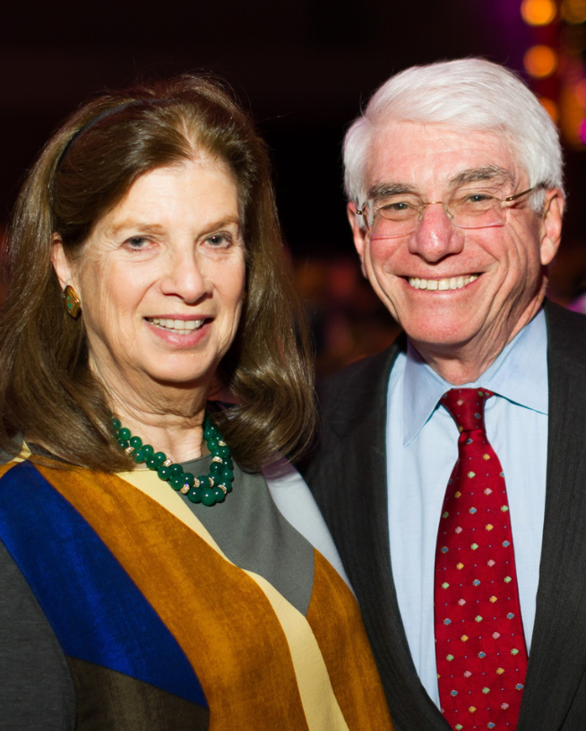 Two smiling older adults, a woman with a colorful scarf and a man in a red tie, standing closely together at an event.