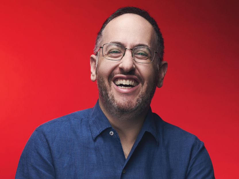 A middle-aged white man wearing glasses and a dark blue button down shirt smiles into the camera in front of a red background.