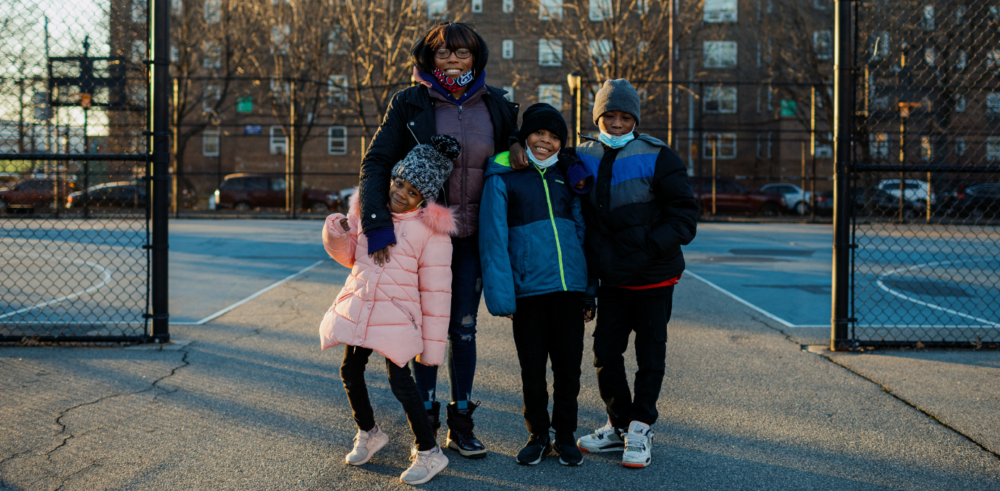 A young Black family including a mother and her three children (two boys and one girl) stands smiling in a playground in front of a brick building.