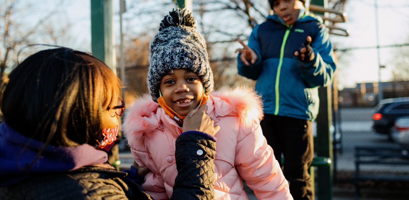 A young Black girl wearing a pink puffer coat smiles at the camera.