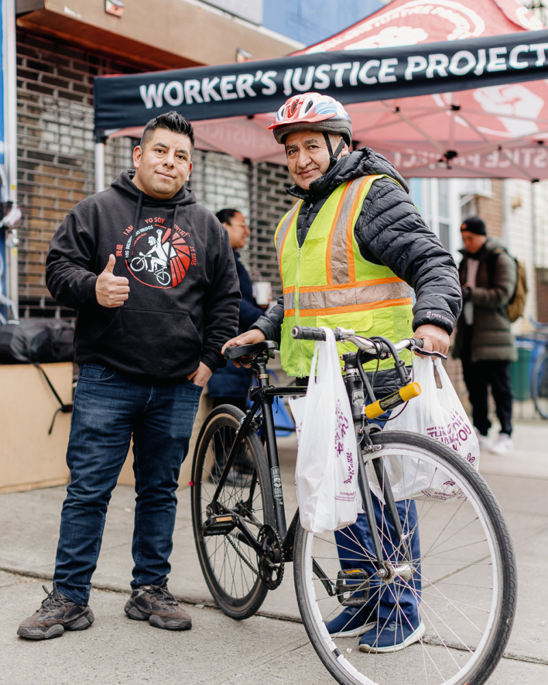 Two men, both standing, are smiling, one is giving a thumbs up while the other is holding up his bicycle