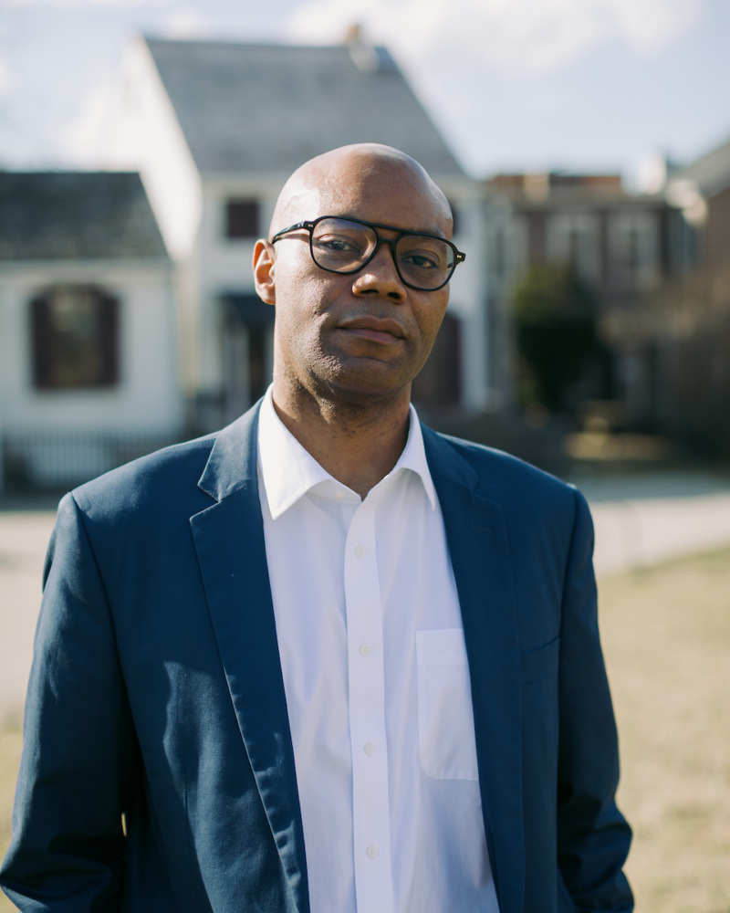 A man wearing glasses, a white shirt, and a blue suit jacket stands outdoors with houses in the background.