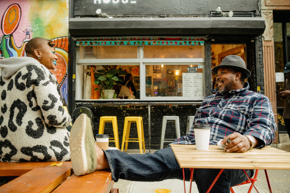Sitting outside a small colorful cafe, a Black man rests his leg on a bench next to a Black woman sitting on the bench facing him wearing a graphic printed coat