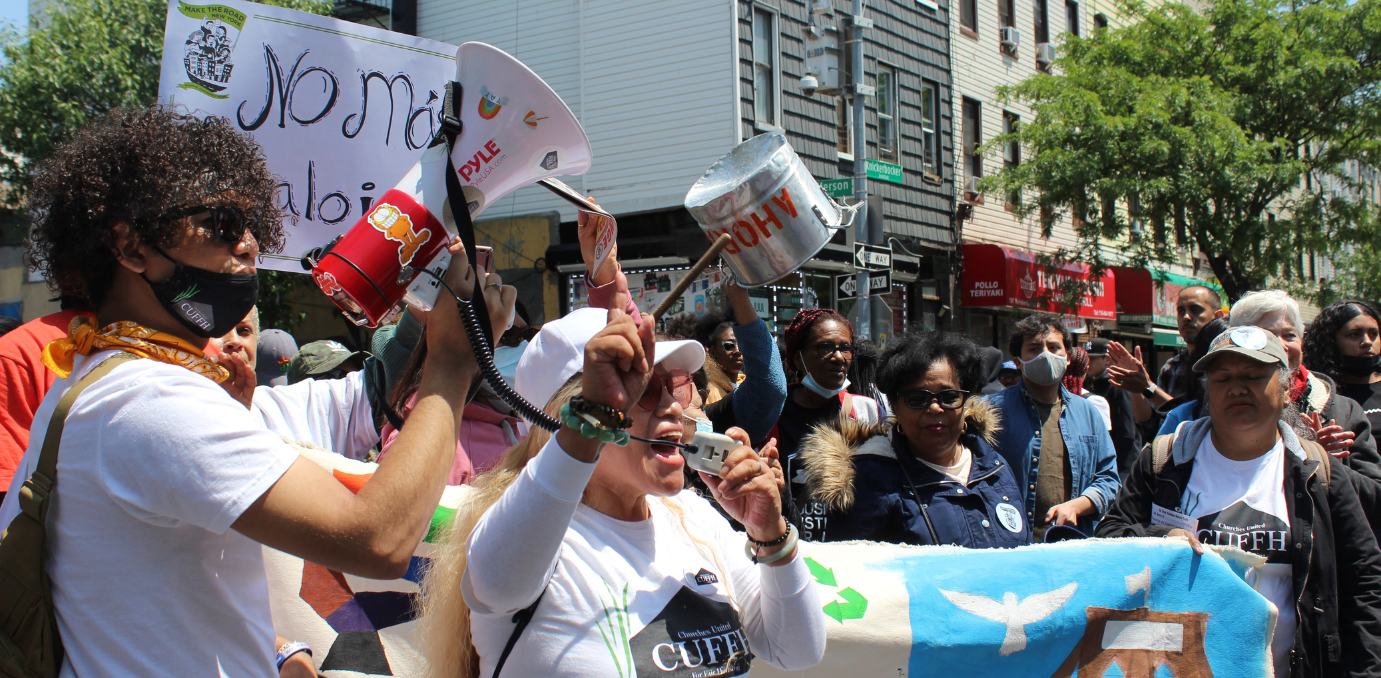 A crowd of organizers gather for a protest. A middle-aged woman wearing a white shirt yells into a bull horn while a young person with curly hair holds the speaker.