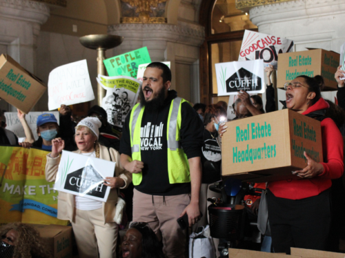A crowd of protesters gather in a government building holding signs that read "Read Estate Headquarters" and "CUFFH." A Latinx man wearing a reflector vest and a VOCAL-NY sweatshirt leads the group in chants.