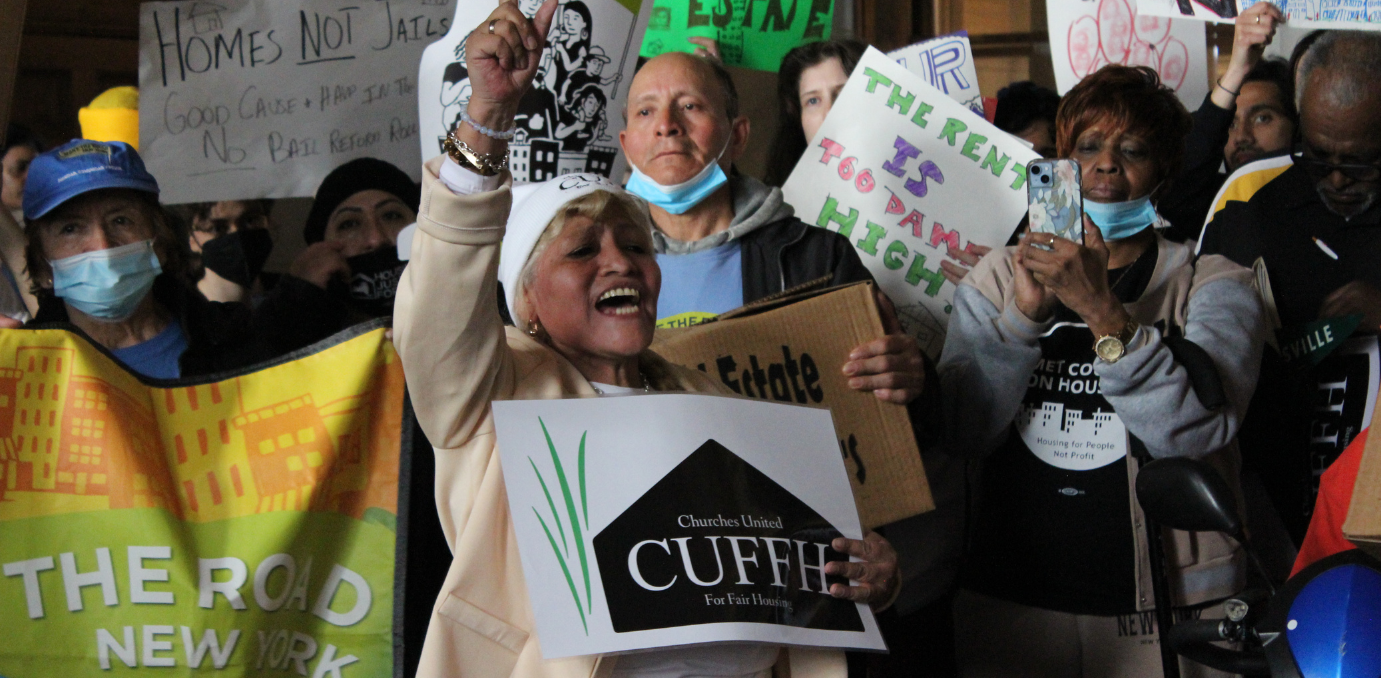 An older Latinx woman holds up her fist passionately while carrying a CUFFH sign. She is wearing a beige sweater and a white beanie.