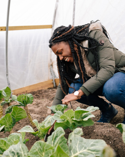 A younger Black woman kneels over a patch of cabbages in a green house