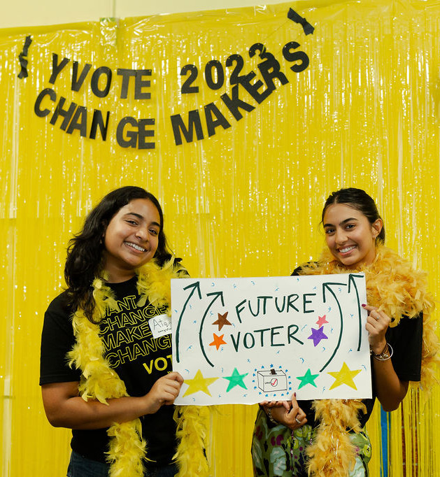 Two young women wearing feather boas smile, posing with a sign that says "Future Voter." They're standing in front of a banner that reads "YVOTE 2023 Changemakers"