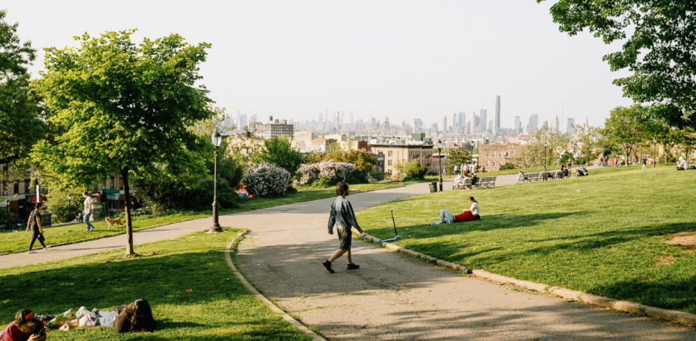 A park on top of a hill shows a path cutting through grass, where a person walks and others sit in the grass. In the background there are trees as a skyline of lower Manhattan.