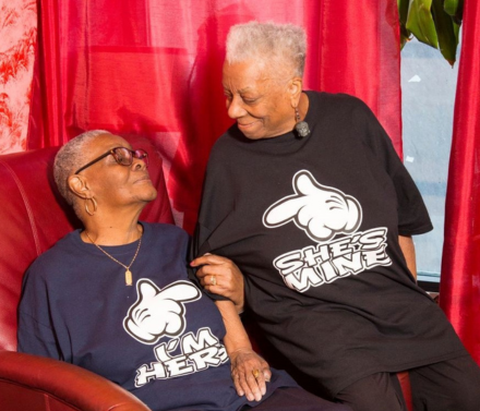 An older Black lesbian couple sitting in an armchair and looking at each other lovingly.