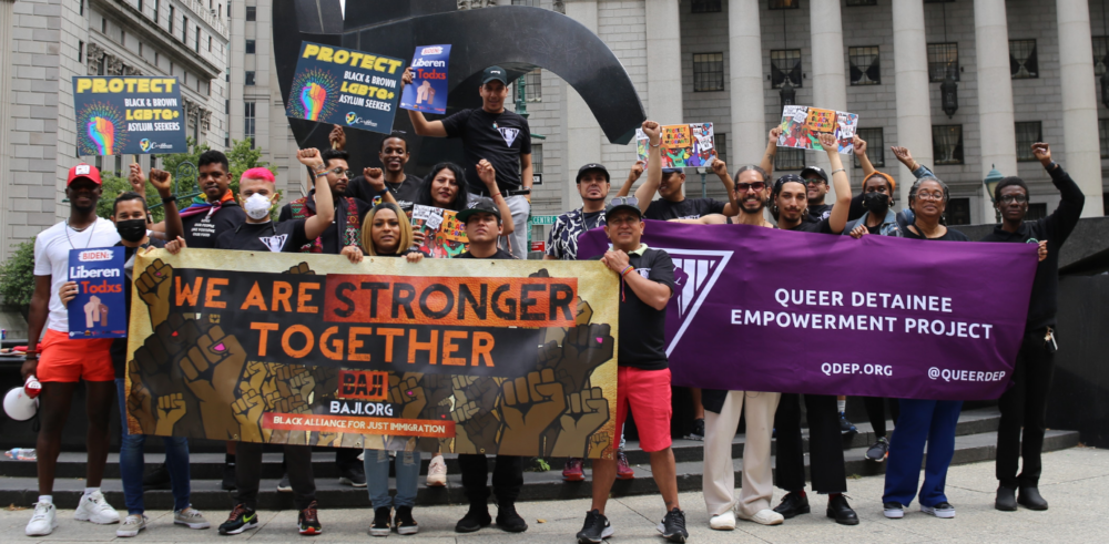 A large crowd of people stand with their fists raised, carrying banners that read "Queer Detainee Empowerment Project" and "We Are Stronger Together."