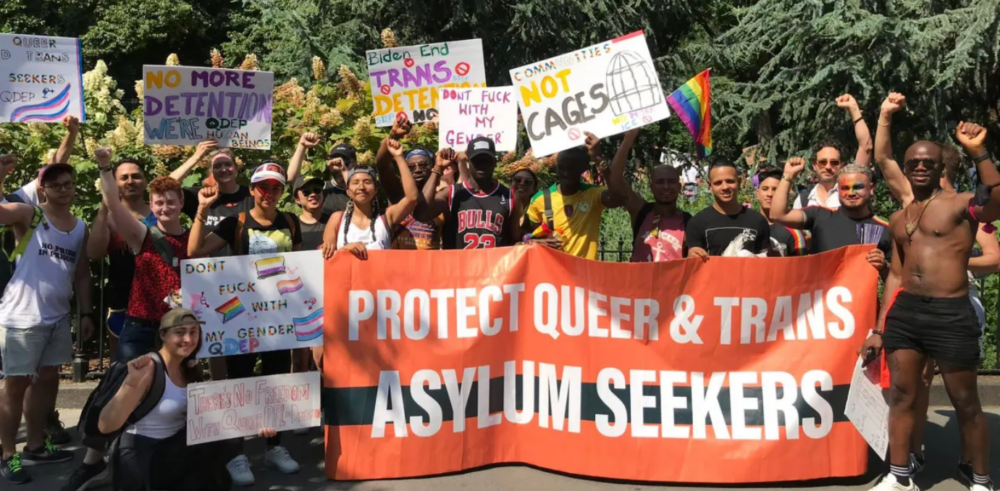 A large crowd cheers and raises their fists behind a large orange banner that reads "Protect Queer & Trans Asylum Seekers."
