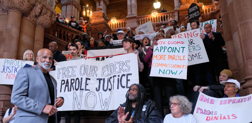 A crowd of people stand on the steps of a government building holding banners that read things like "Free Our Elders Parole Justice Now"