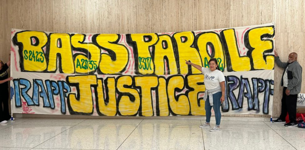 A middle-aged woman wearing a white tshirt points to a large banner which says "Pass Parole Justice" in dark yellow letters.