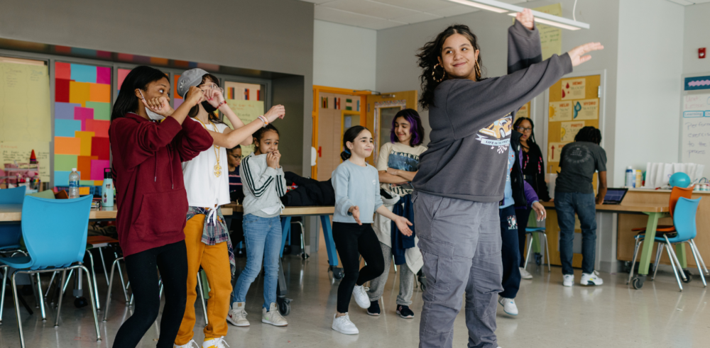 A young woman wearing a gray sweatsuit leads a group of younger girls in a dance routine in a classroom, with their arms in the air.