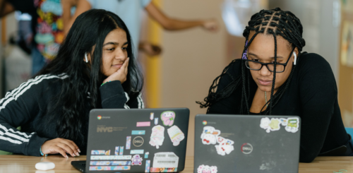 Two young girls of color wearing black sweatshirts sit next to each other and work on their laptops.