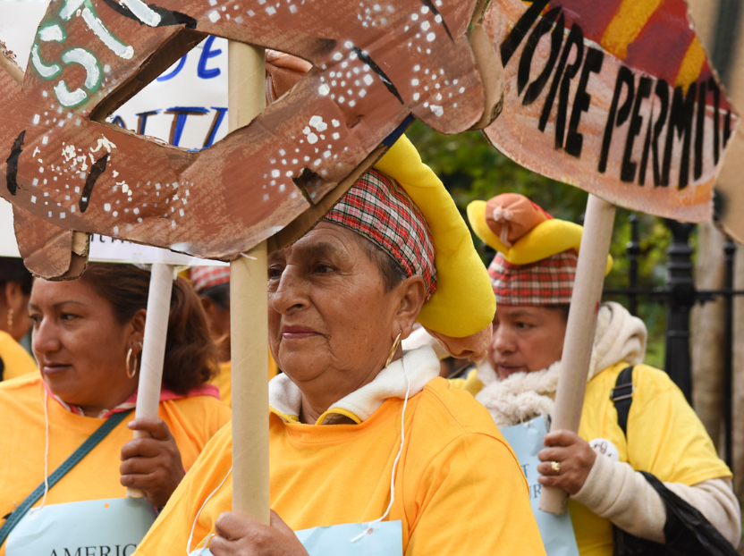An older Latinx woman wearing a dark yellow shirt and holds a sign shaped into a pretzel that is slightly out of frame.