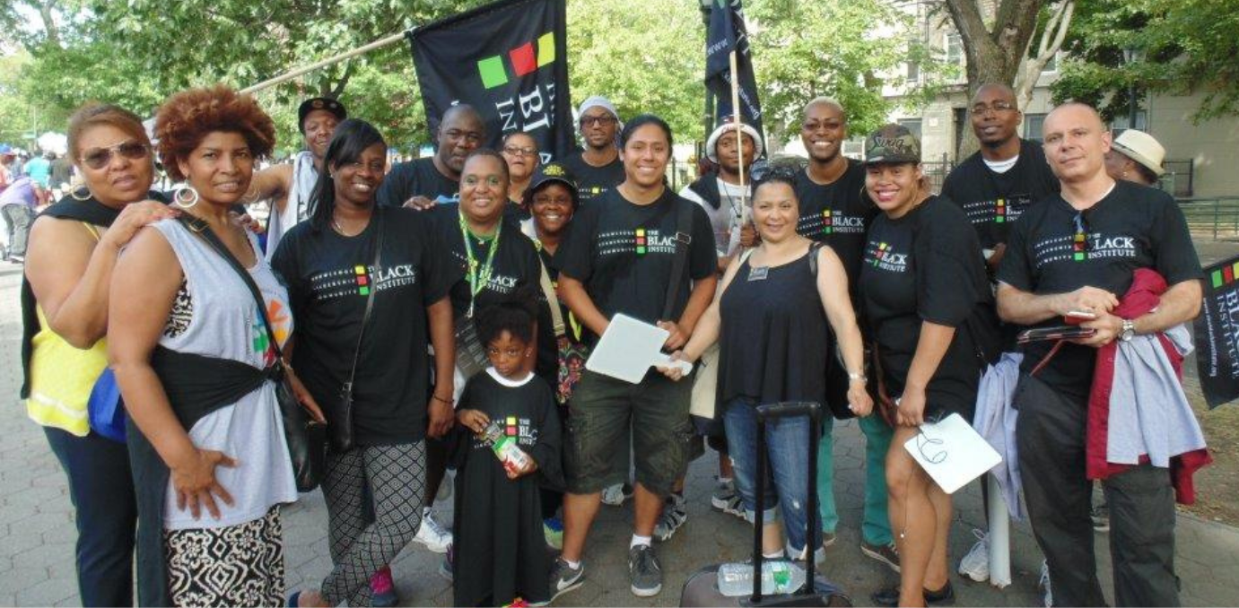 A crowd of people wearing black "The Black Institute" tshirts stand together smiling at the camera. A The Black Institute flag waves in the background.