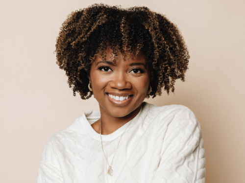 A young Black woman with an afro wearing a white sweater smiles in front of a beige background.