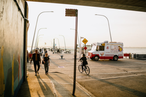 A group of people walking down a sidewalk near an ice cream truck on a pier in Bay Ridge, Brooklyn near sunset.