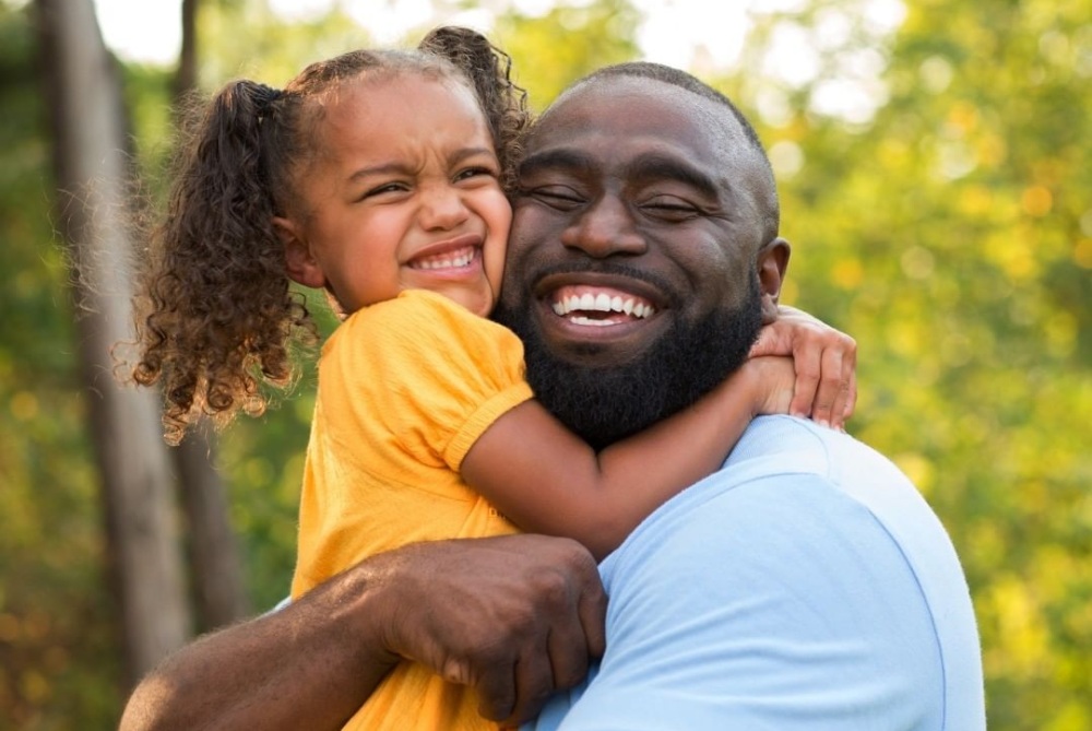 A Black man hugs his daughter in the park.