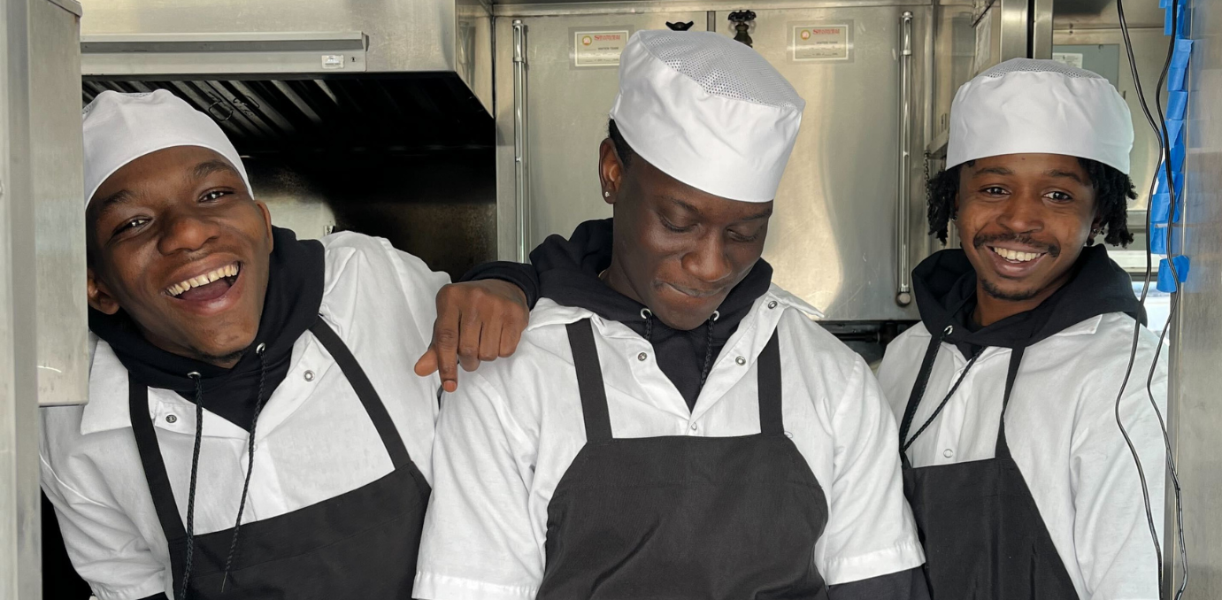 Three men in aprons and hats smiling in a kitchen.
