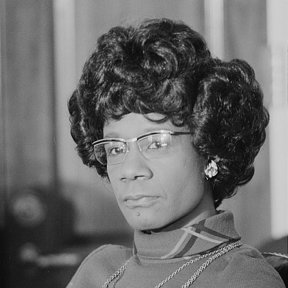 A black and white photo of Congresswoman Shirley Chisholm. She is a Black woman with curly hair and glasses.