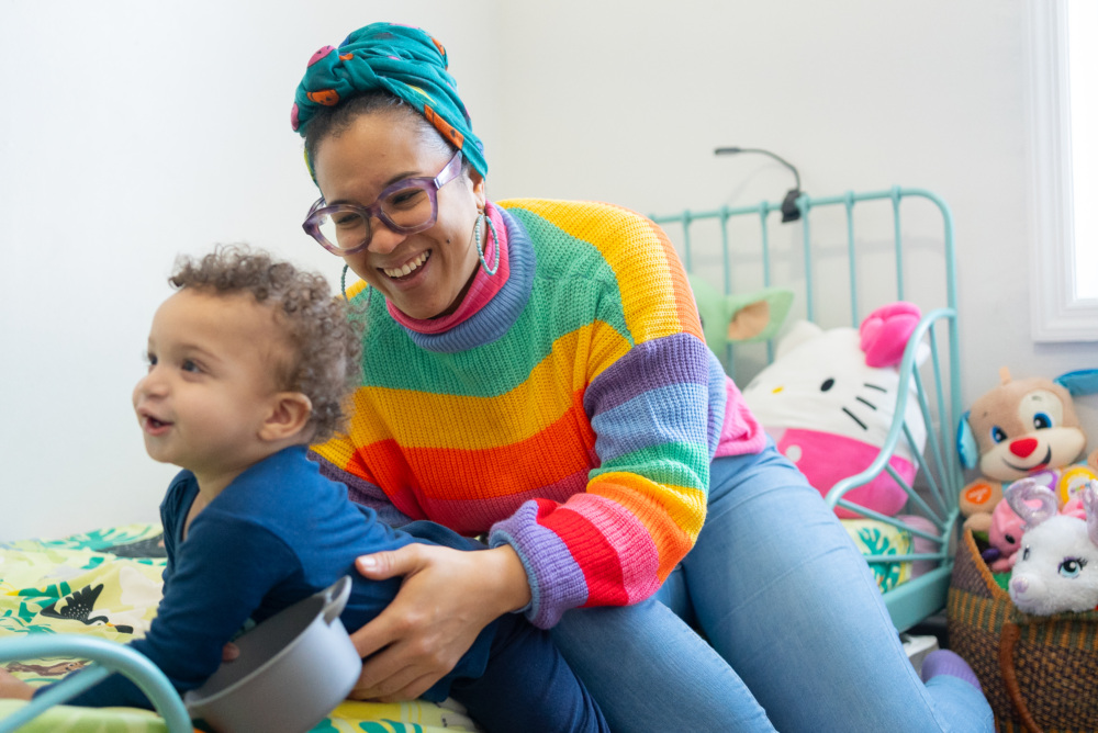 Adult in a colorful sweater and headwrap plays with a child in a bedroom decorated with plush toys and a small bed.