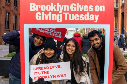 Four people smiling through a large frame with "Brooklyn Gives on Giving Tuesday" text, holding signs on the street in DUMBO, Brooklyn.