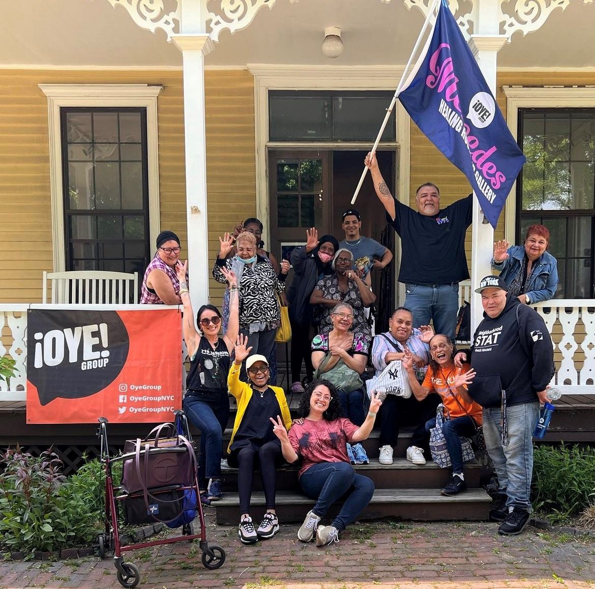 A group of older adults posing on the steps of a house. One person is holding up a large blue and pink flag that says Oye Mercedes Healing Room Gallery.