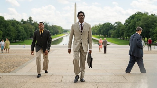 Two men in suits walking in front of the washington monument.