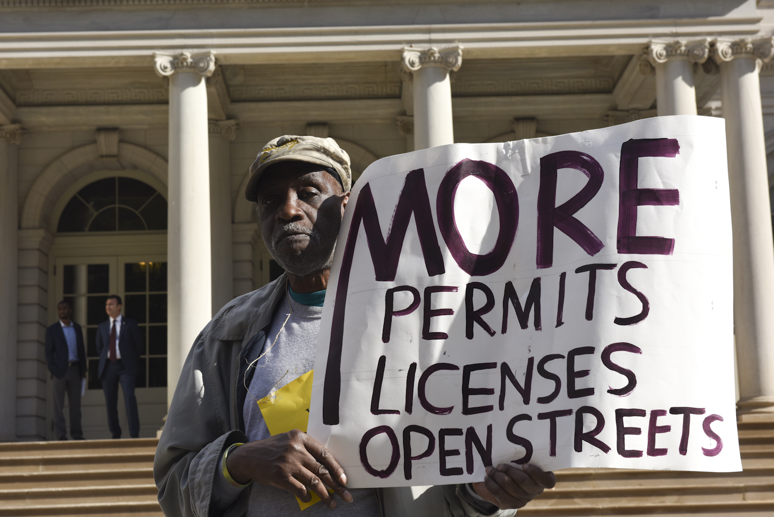 A Black man stands in front of New York CIty Hall holding a sign that says more permits licenses open streets.