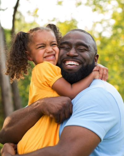 A Black man hugs his daughter in the park.