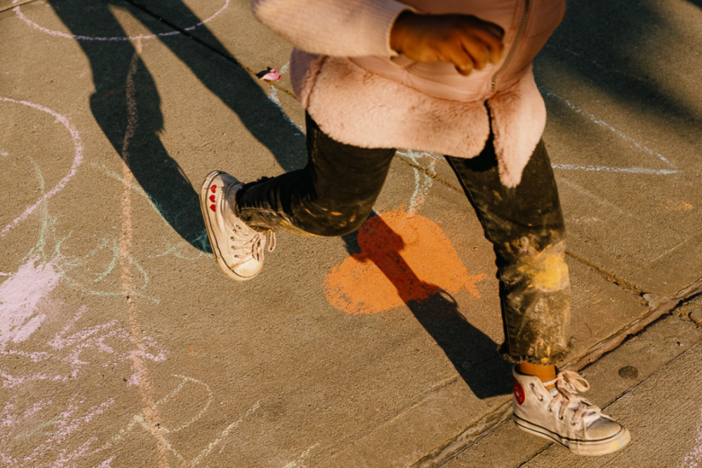 A girl in a pink jacket is running on a sidewalk with chalk.