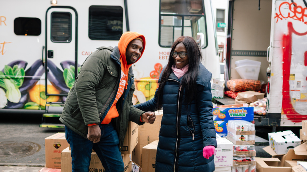 Two food pantry staff, a Black man and woman, standing in front of a truck with boxes of food and groceries.