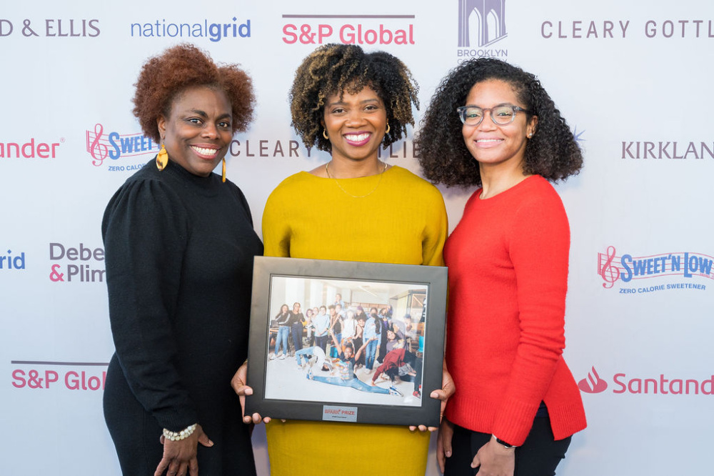 Three Black women pose with a framed photo on a red carpet.