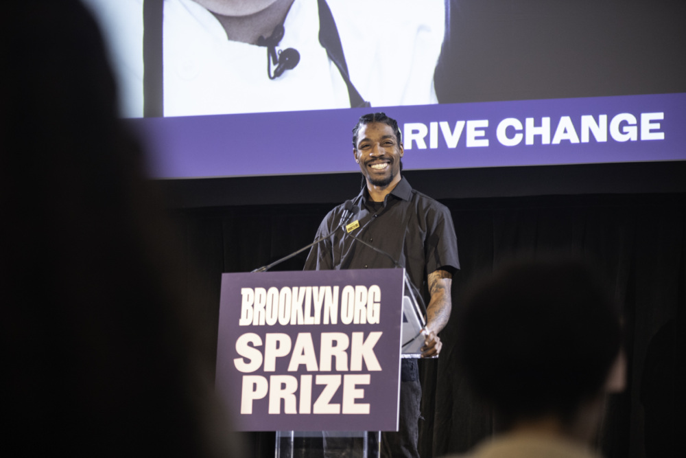A Black man is standing at a lectern with a microphone, smiling as he addresses an audience, with a sign reading "Brooklyn Org Spark Prize" visible on the front of the lectern.