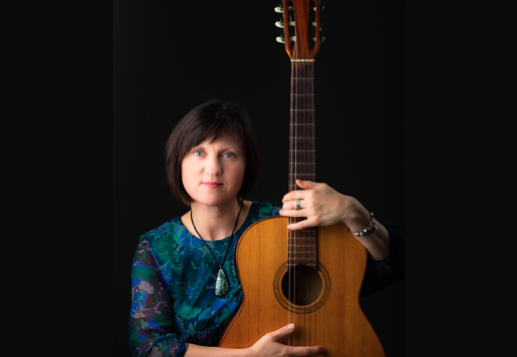 A woman holding an acoustic guitar in front of a black background.