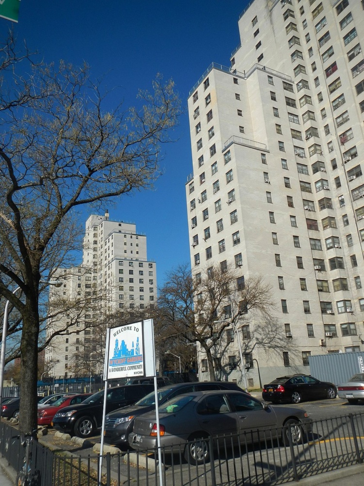 Tall residential buildings rising above a welcoming sign with parked cars and leafless trees on a clear day.