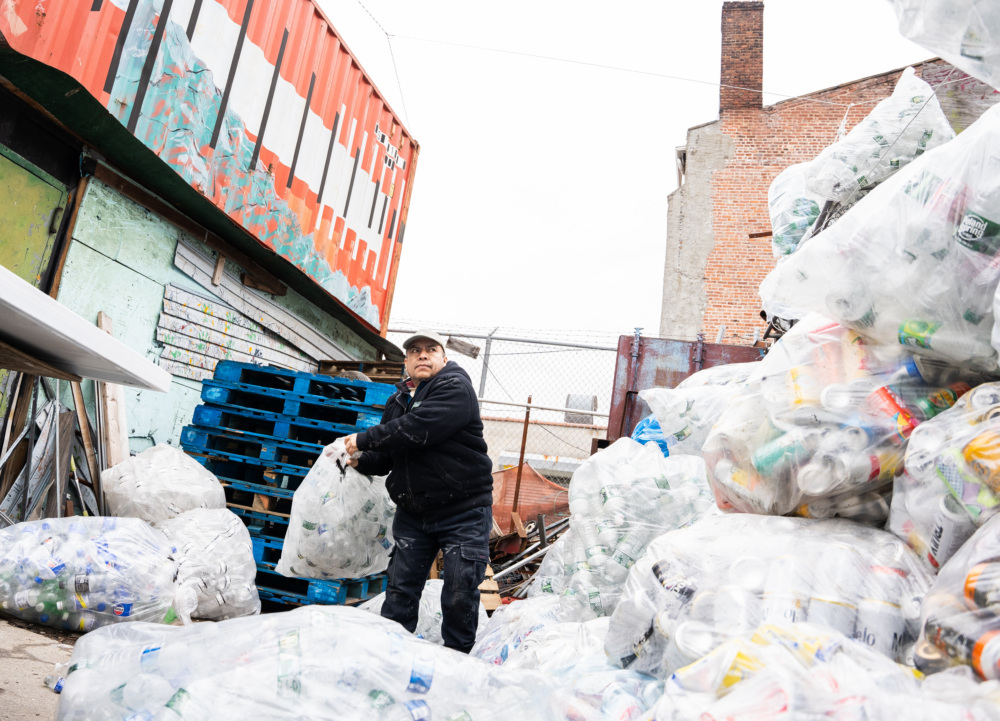 Worker handling large bags of recyclable materials in an outdoor recycling facility.