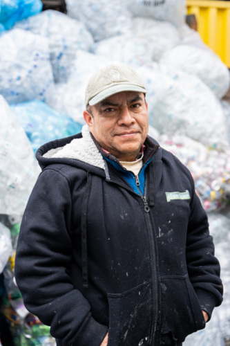 A man wearing a cap and a black jacket stands in front of large bags of recyclable materials.