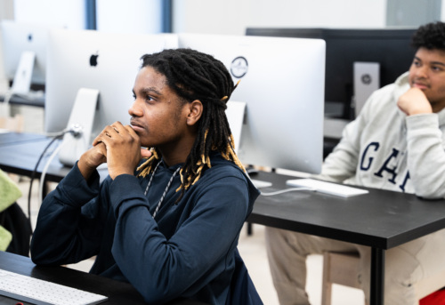 Two young Black men sit at a table in a computer lab, one with attentive focus towards the front, the other appears more relaxed, with computers in the background.