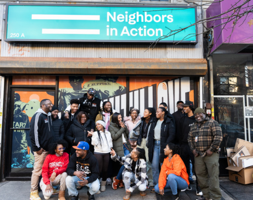 A diverse group of people gathers smiling in front of the "neighbors in action" office, suggesting a sense of community and teamwork.