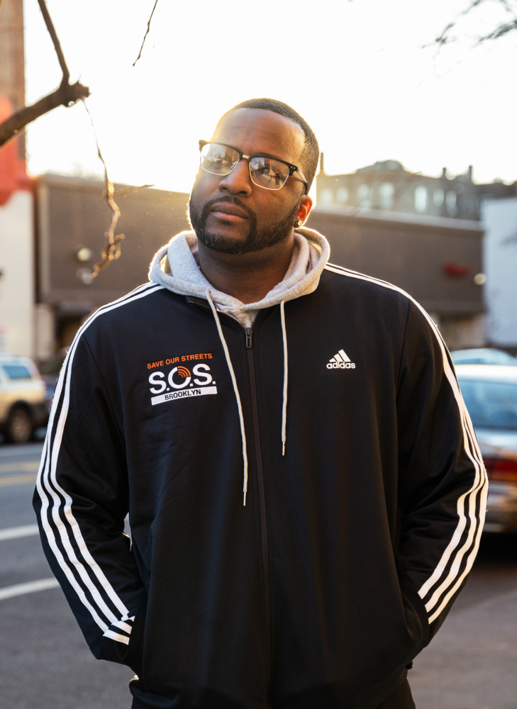 A Black man in a black adidas jacket and glasses standing outdoors during golden hour.