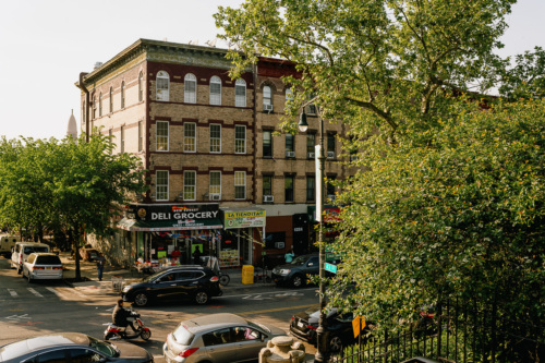 Old brick buildings with a street-level deli grocery store on a sunny day in a bustling urban neighborhood.