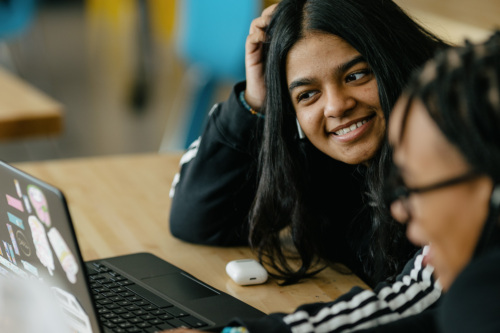 A smiling young woman sitting at a table with a laptop and another person partly in view.