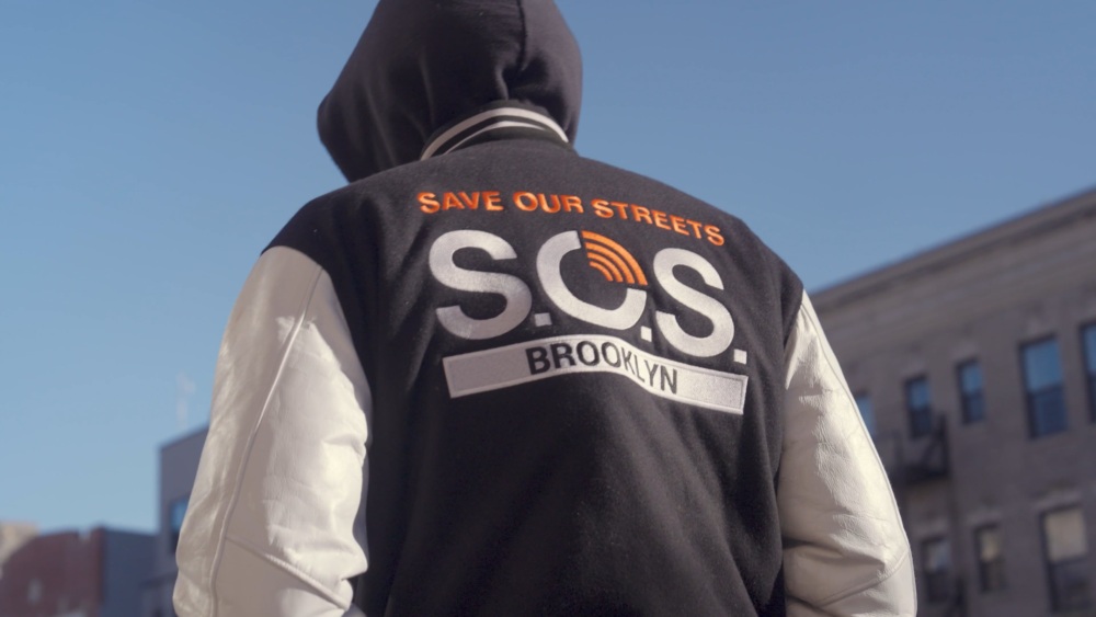 A person seen from behind wearing a black hoodie with a logo that reads "save our streets s.o.s. brooklyn.