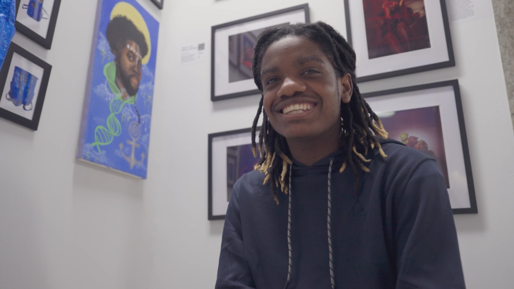 A young person with shoulder-length dreadlocks and a black outfit smiling in an art gallery with various framed works on the walls.
